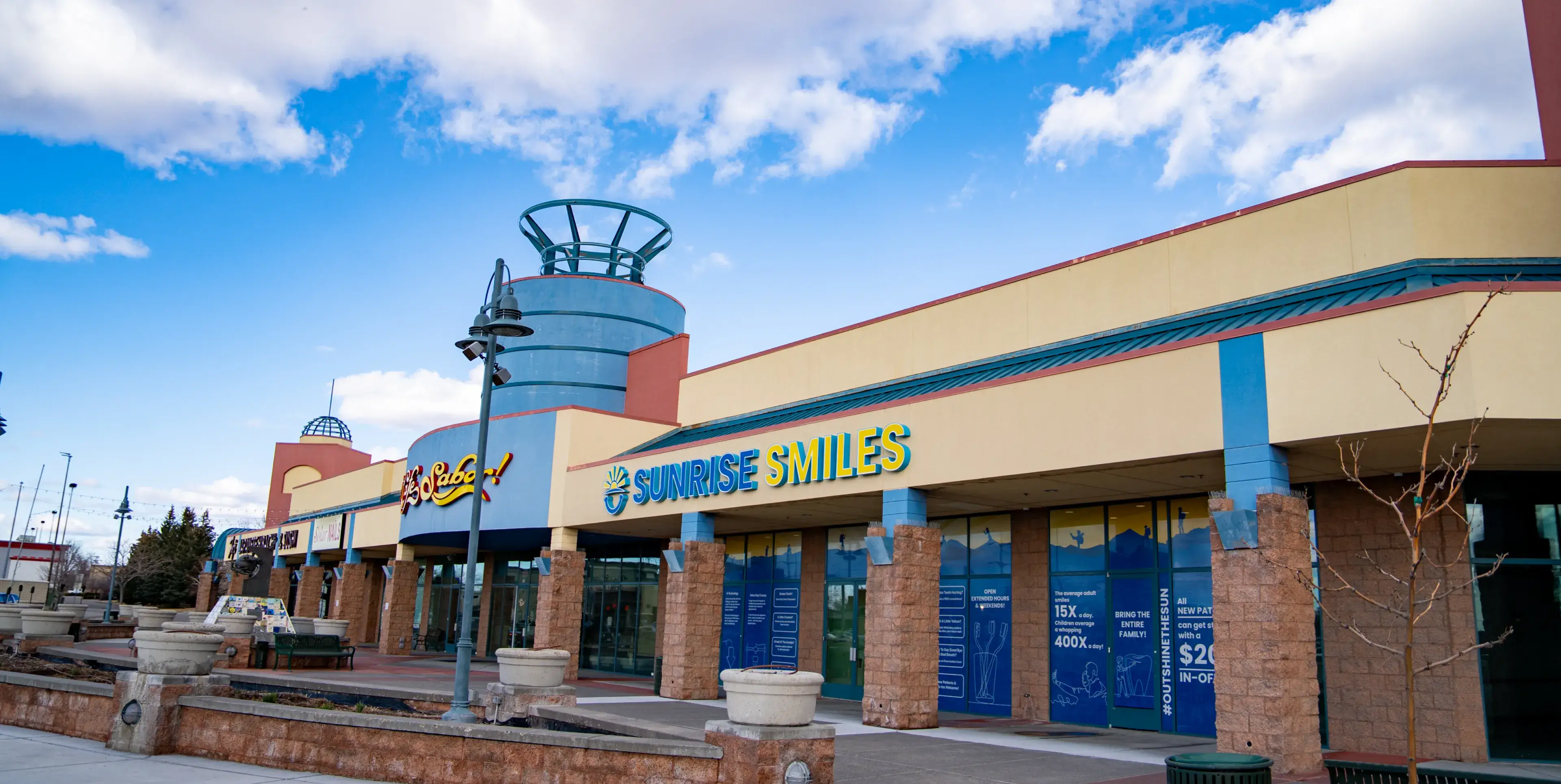 Exterior of a commercial plaza with storefronts including Sunrise Smiles dental office and a café under a blue sky with scattered clouds.