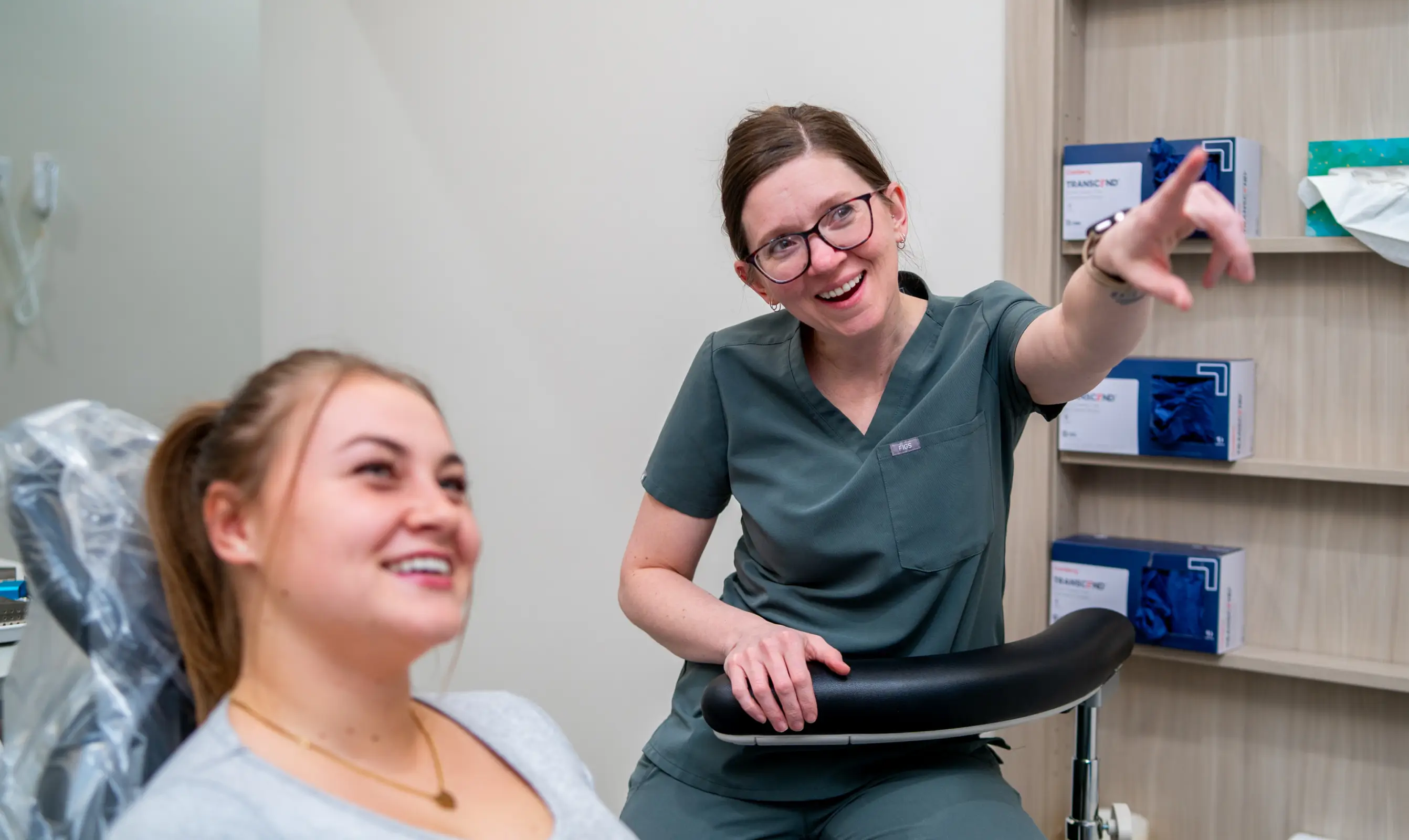 Female healthcare worker in scrubs smiling and pointing while talking to a seated female patient.