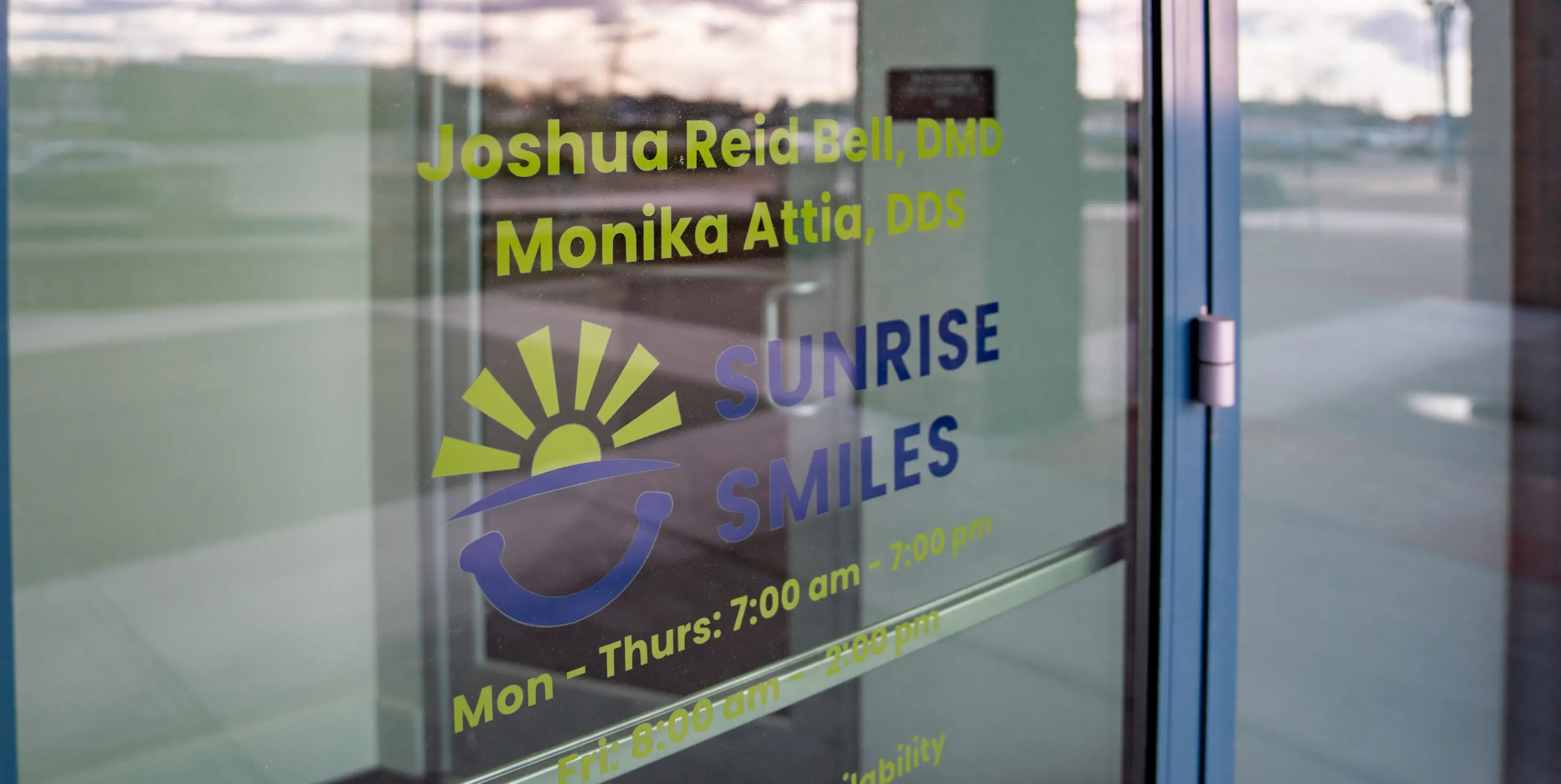 Glass door showing the Sunrise Smiles dental office hours and names of dentists Joshua Reid Bell, DMD, and Monika Attia, DDS.