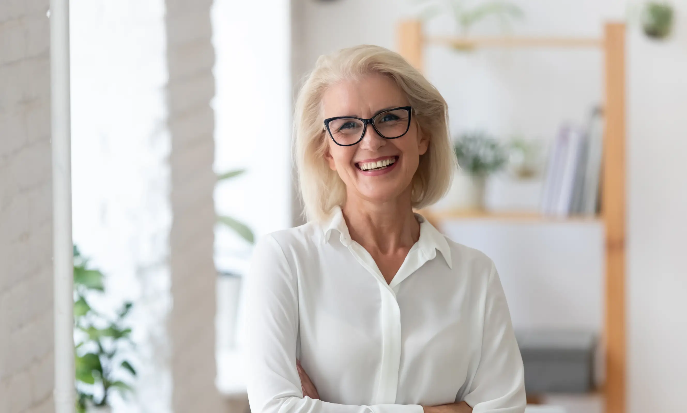 Smiling woman wearing glasses and white shirt standing with arms crossed indoors.
