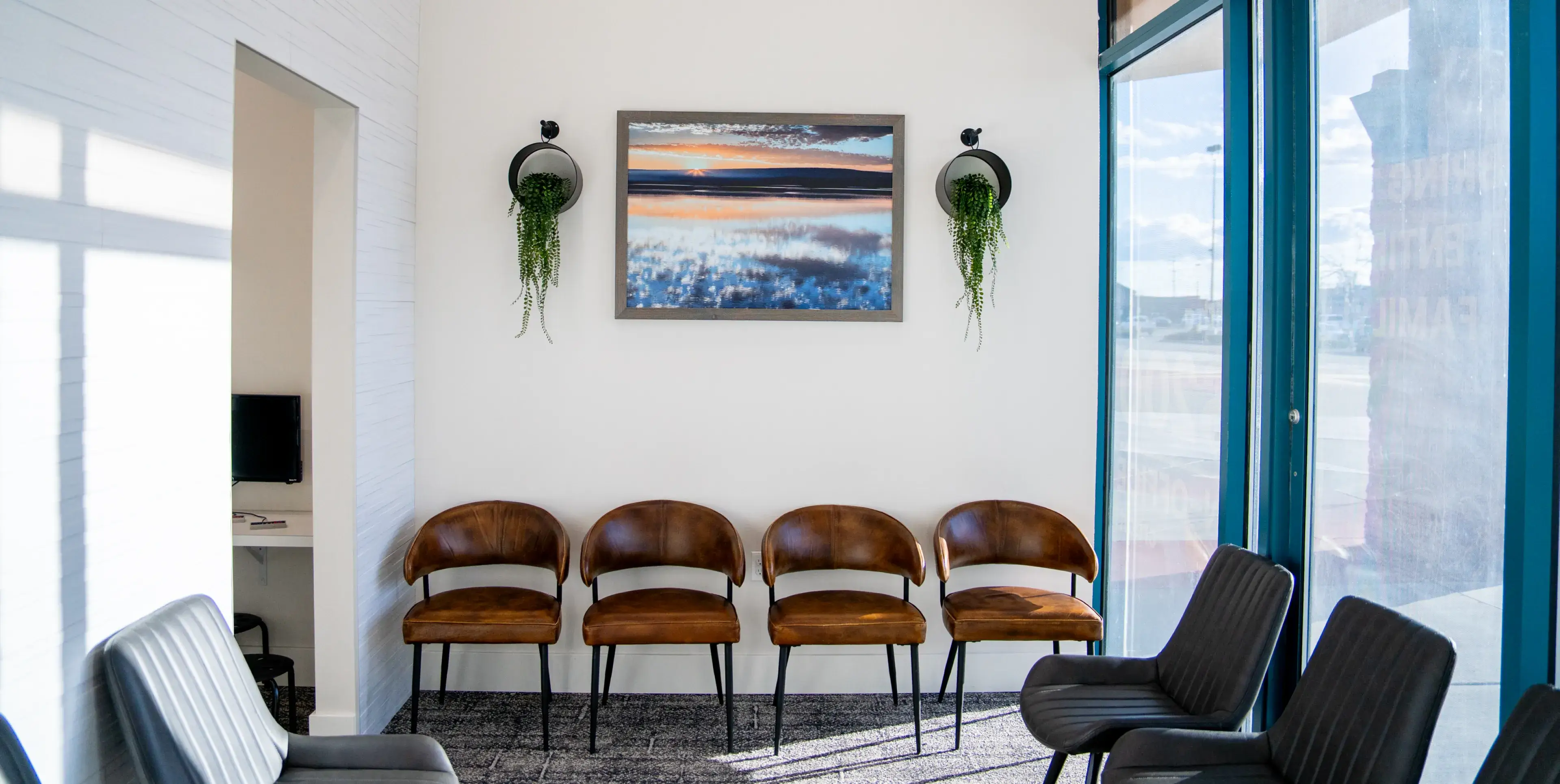 Modern waiting area with four brown leather chairs, two black chairs, a large window, wall-mounted plants, and a framed landscape photograph.