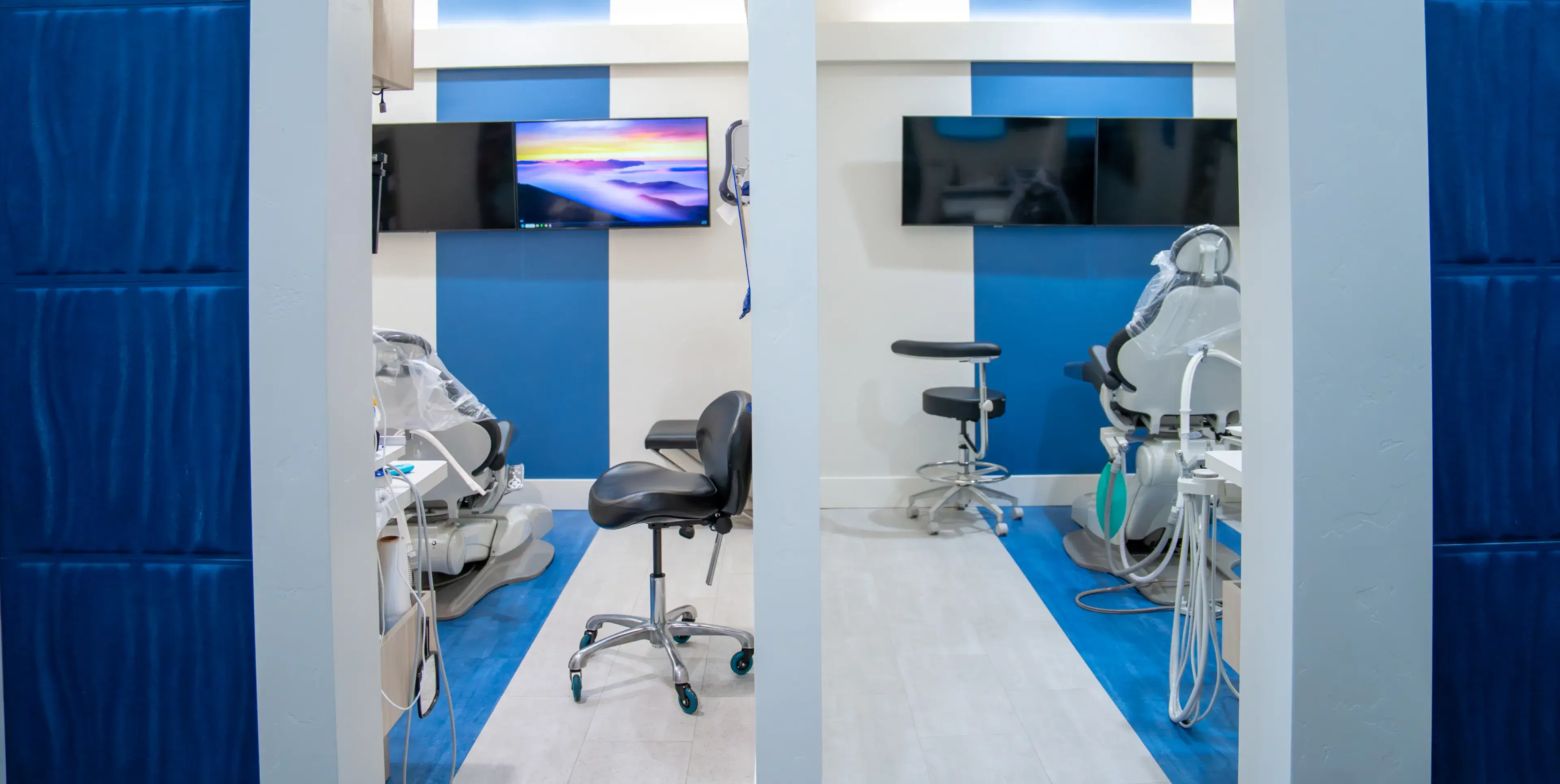Modern dental clinic treatment rooms with blue and white walls, dental chairs, stools, and wall-mounted monitors.