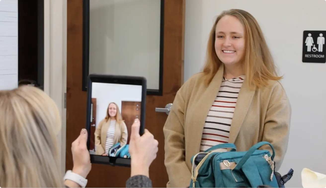 Woman smiling as another person takes her photo with a tablet in an indoor setting.